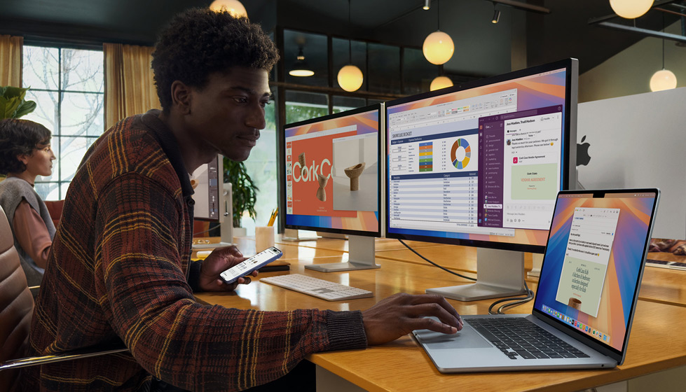 Eine Person in einer Büroumgebung nutzt ein aufgeklapptes MacBook Air mit zwei externen Displays und Magic Keyboard. In der anderen Hand hält sie ein iPhone.