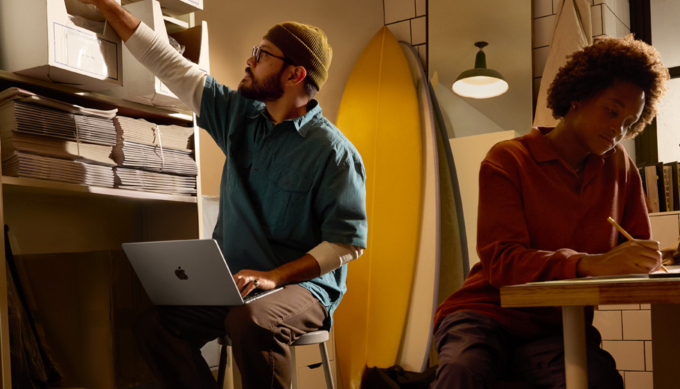 Person in einer Arbeitsumgebung, die auf einem Stuhl sitzt und ein MacBook Air auf ihrem Schoss nutzt, das nicht am Strom angeschlossen ist. Mit der anderen Hand greift sie nach einer Schachtel auf einem hohen Regal.